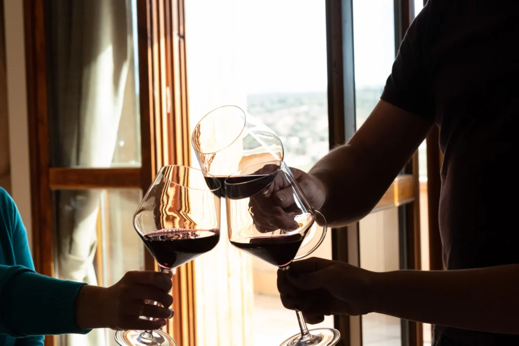 Tres personas brindando con copas de vino tinto junto a una ventana iluminada. Esta imagen se utiliza para ilustrar una entrada sobre cómo catar un vino.